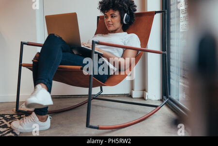 Young african woman sitting relaxed on chair wearing headphones and working on laptop. Female software programmer working at tech startup. Stock Photo