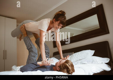 Happy couple playing in bed and having fun. Man lying on bed and lifting his wife with his feet holding her hands. Stock Photo