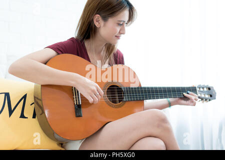 Young beautiful Asian woman sitting on sofa playing guitar - girl music, having fun concept Stock Photo