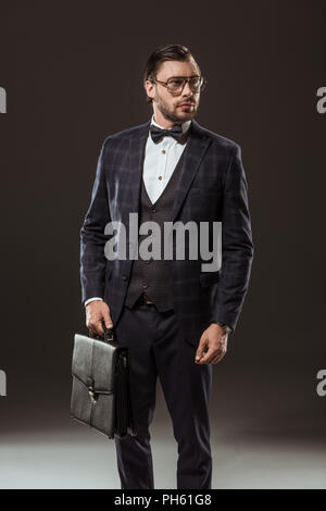 handsome buisnessman in suit, eyeglasses and bow tie holding briefcase and looking away on black Stock Photo