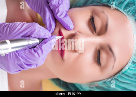 Procedure of professional apply permanent make up in beauty salon on woman lips. Girl with protective green cap lies down in bed and having tattoo Stock Photo