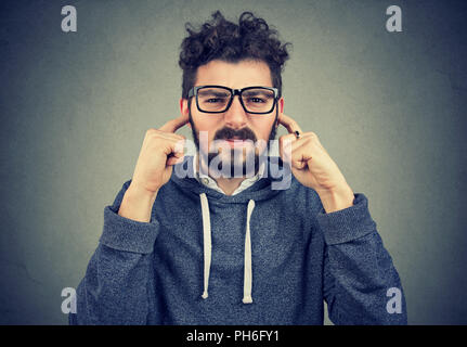 Annoyed young hipster man looking grumpy at camera and covering ears from noise avoiding loud noise and conflict situation on gray background Stock Photo