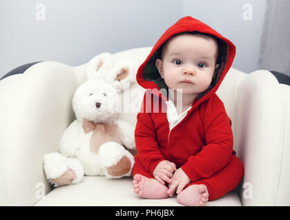 Portrait of cute adorable Caucasian smiling sad baby boy girl with black brown eyes in red hoodie shirt sitting in chair with toy looking directly in  Stock Photo