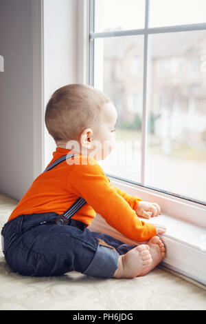 Portrait of cute adorable Caucasian baby boy with black eyes in orange shirt onesie, jeans with suspenders barefoot sitting on windowsill looking away Stock Photo