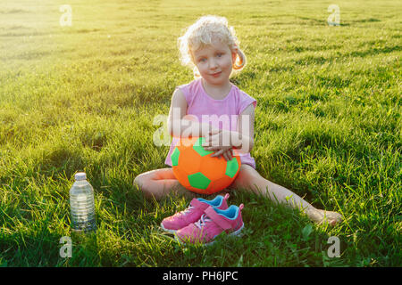 Portrait of happy cute adorable white Caucasian girl holding soft soccer ball. Child sitting in grass outside after playing soccer football game on su Stock Photo