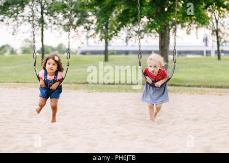 Portrait of two happy smiling little toddlers girls friends swinging on swings at playground outside on summer day. Happy childhood lifestyle concept. Stock Photo