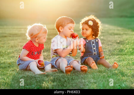 Group portrait of three white Caucasian and latin hispanic children boy and girls sitting together sharing and eating apple food. Love friendship chil Stock Photo