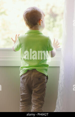Portrait of white Caucasian child boy at home looking out of window outside. View from back. Kid waiting for someone Stock Photo