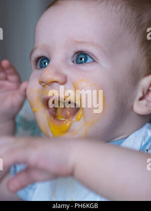 Messy baby boy with blue eyes eating lunch and smiling Stock Photo