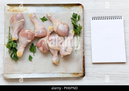 Uncooked raw chicken legs on a baking sheet paper on tray and blank notepad, overhead view. White wooden background. Space for text. Stock Photo