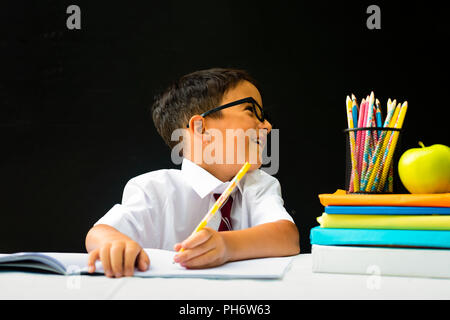 Cute smart handsome school boy in white shirt with eye glasses, writing at the desk in classroom, distracted, talking happy first grader pupil, smilin Stock Photo