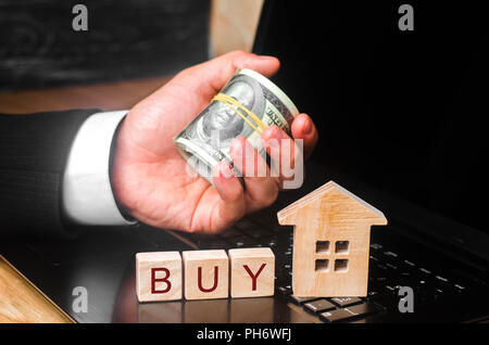 The realtor's hand holds a bundle of money behind the miniature house. The concept of buying and selling real estate, investment. Purchase of building Stock Photo