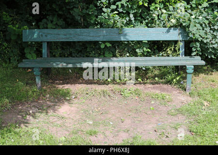 Green painted traditional wooden park seat on a metal frame in a shady corner overgrown by adjacent shrubs with a worn muddy area of grass lawn in the Stock Photo