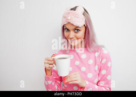 Attractive girl in pink pajamas and sleep mask enjoying her morning coffee. Portrait of young woman on white background with white mug Stock Photo