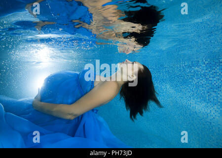 A pregnant woman in a blue dress looks at her reflection by the underwater in the pool Stock Photo