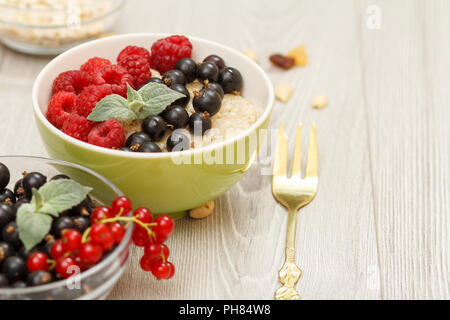 Oatmeal porridge in porcelain bowl with currant berries and raspberries, decorated with mint leaves. Glass bowls with oat flakes and currant berries.  Stock Photo