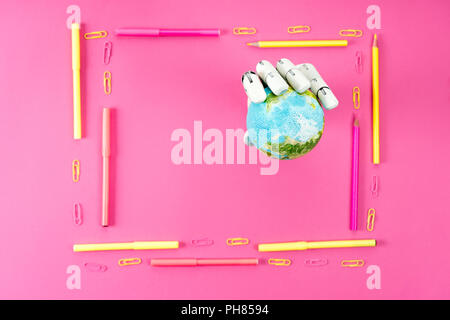 top view of robotic hand holding globe inside of frame made of pencils and markers on pink Stock Photo
