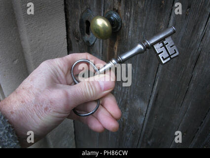 Large metal church door key held in a man's hand. The keyhole, doorknob and door can be seen. Stock Photo