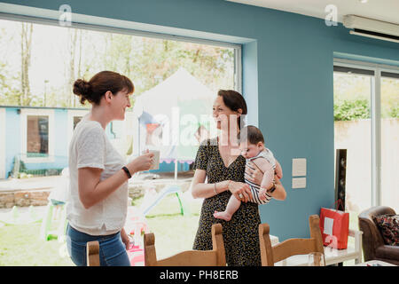 Two mid adult women standing in the kicthen at home and socialising. One woman is holding her baby son. They are talking and laughing together. Stock Photo