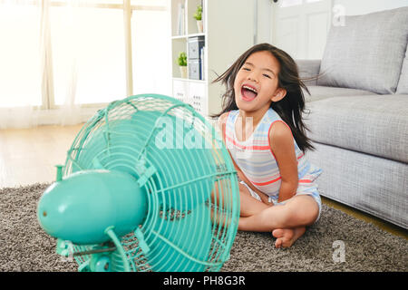 litle cute girl laughing and sitting in front of fan. refreshing herself from hot weather with legs crossed. Stock Photo