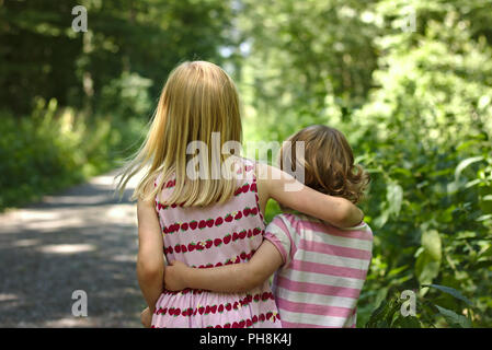 Two little girls wearing summer dresses walk arm in arm along a sunny forest trail Stock Photo