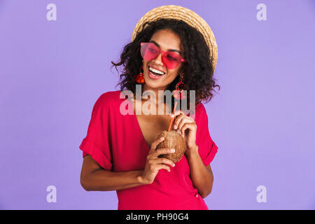 Happy african woman in dress, straw hat and sunglasses posing with cocktail while looking away over purple background Stock Photo
