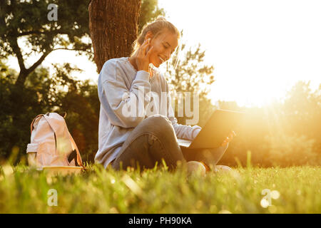 Portrait of a cheerful young girl in earphones with backpack sitting with legs crossed on a grass at the park, using laptop computer Stock Photo