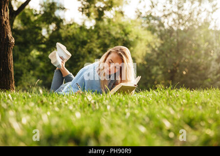 Portrait of a lovely young girl laying on a grass at the park, reading a book, taking notes Stock Photo