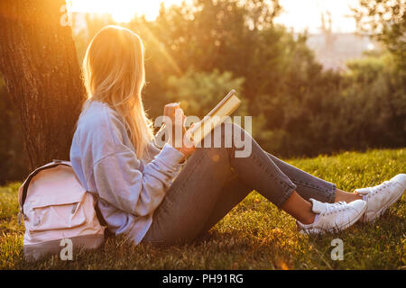 Portrait of a young girl sitting on a grass at the park, reading a book, taking notes Stock Photo