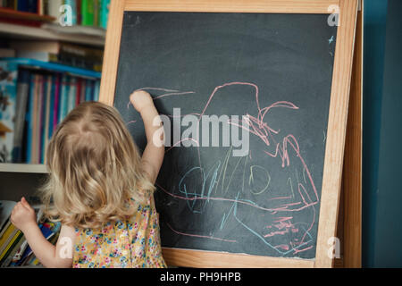 Rear view of a little girl drawing and writing on a blackboard. The little girl is reaching up and drawing, using different coloured chalk on the blac Stock Photo