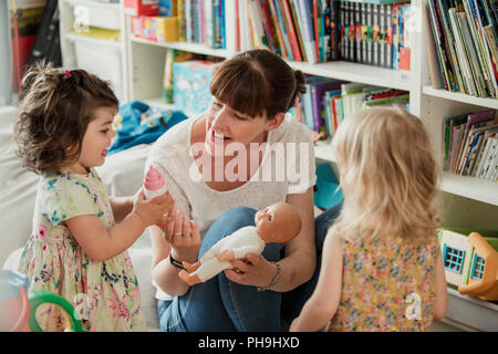 Mother and her two daughters are looking at a toy baby doll and talking about giving it a bottle. The mother is holding the doll baby and passing her  Stock Photo