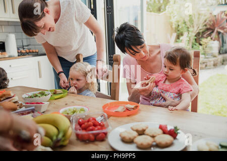 Two mid adult mothers giving their daughters new food to try. They are in the kitchen enjoying their healthy lunch. Stock Photo