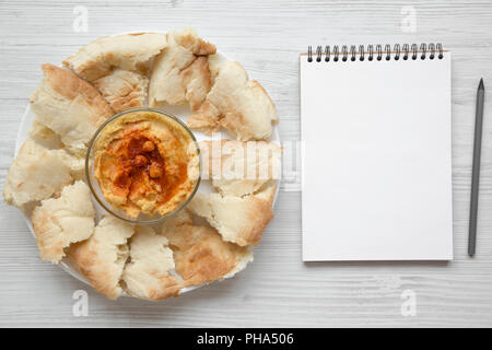 Bowl of Hummus with chickpeas, paprika, olive oil and pita bread on white plate. Blank notebook over white wooden background, view from above. Top vie Stock Photo