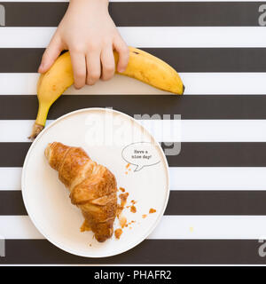 Child's hand taking organic banana from black and white stripes background. top view. French croissant served on white plate with sign Have a nice day Stock Photo