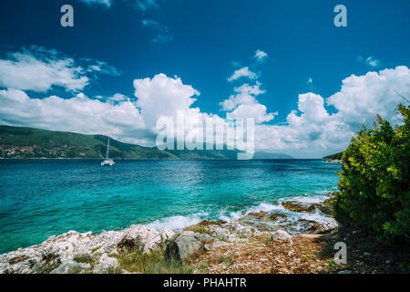 Crystal clear transparent blue turquoise teal Mediterranean sea water in Fiskardo town. White yacht in open sea at anchor under amazing white clouds, Kefalonia, Ionian islands, Greece Stock Photo