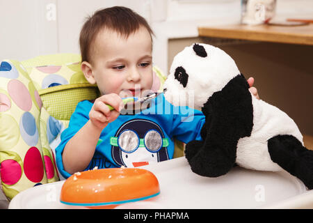 Close-up portrait of cute adorable little boy toddler two years old sitting in high chair in kitchen eating meal lunch dinner using fork and feeding h Stock Photo