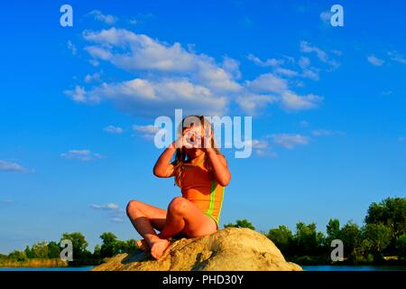 Beautiful girl sitting on a big rock. Little girl is wearing swimsuit. Girl is making heart shape gesture by her hands. Summer and happy childhood concept. Copy space in bright blue sky Stock Photo