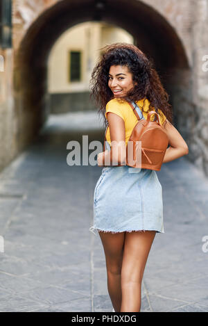 Rear view of smiling black tourist woman with curly hair walking on the street. Arab traveler girl in casual clothes in the street. Happy female weari Stock Photo