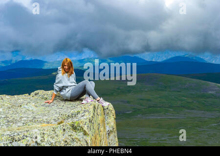 The red-haired girl sits alone on the edge of a cliff in the rays of light on a rock and is afraid to look down by turning her head, Altai mountains a Stock Photo