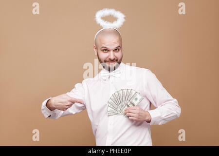 Happy handsome middle aged bald angel in shirt and white halo standing, holding and pointing at many dollars and looking at camera with smile. indoor  Stock Photo