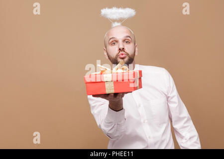Happy handsome middle aged bald bearded angel in shirt and white halo on head standing holding red gift, looking at camera and sending kiss. indoor st Stock Photo