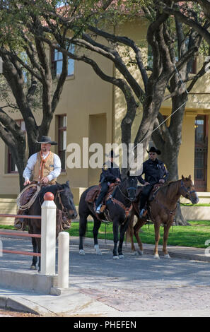 A Texan Cowboy gets ready to herd steer while two Texan mounted police look on in Fort Worth Stockyards, Texas. Trees and buildings in  background.