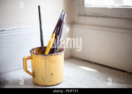 Small plastic bucket with used artist paint brushes on a corner shelf below a window Stock Photo