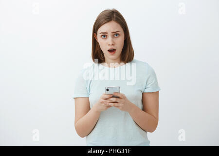 Worried nervous girl asking advice from friend trying text guy she loves standing concerned and troubled over white wall holding smartphone staring unsure and perplexed at camera with opened mouth Stock Photo