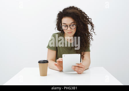 Checking schedule while waiting for client. Portrait of good-looking confident female creative coworker in glasses, sitting at table, drinking coffee and using digital tablet to communicate with boss Stock Photo