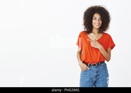 Wanna go there together. Portrait of happy confident and friendly woman with dark skin and afro haircut, holding hand in pocket and pointing left with index finger, standing over grey wall Stock Photo