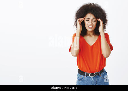 Poor girl having terrible headache. Studio shot of upset european female with afro haircut, touching temples and frowning, grimacing from pain, suffering migraine and standing unfocused Stock Photo
