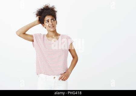 Indoor shot of unsure good-looking curly-haired woman in striped t-shirt, scratching back of head and frowning while looking up with confused unaware expression, feeling doubt or hesitating Stock Photo
