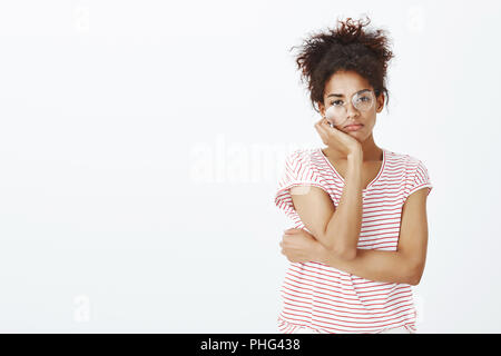 Studio shot of bored indifferent cute daughter in striped t-shirt and glasses, looking with careless expression at camera, leaning head on hands, being unimpressed and having no care over gray wall Stock Photo