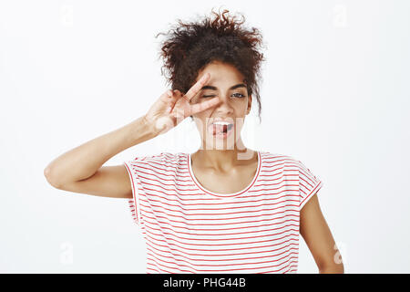 Close-up shot of ougoing confident young woman in striped t-shirt, showing victory or peace gesture over eye, winking and sticking out tongue joyfully, standing carefree over gray background Stock Photo